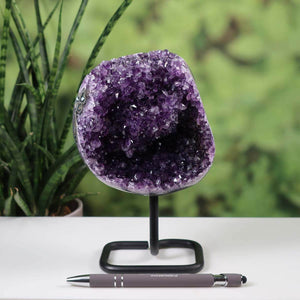 Deep purple Uruguay Amethyst geode on metal stand displayed on a white table with a pen for scale and green plants in the background.