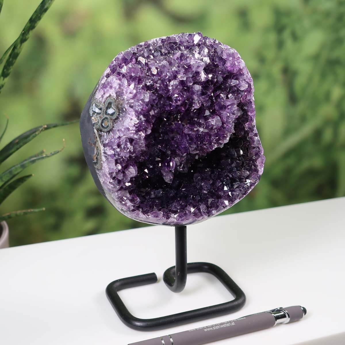 Deep purple Uruguay Amethyst geode on metal stand displayed on a white table with a pen for scale and green plants in the background.