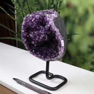 Dark purple Uruguay amethyst geode on metal stand displayed on a white surface with a pen for scale and green foliage in the background.