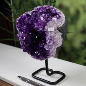 Dark purple Uruguay amethyst druse on black metal stand next to a pen, showcasing intense sparkle and natural crystal formation.