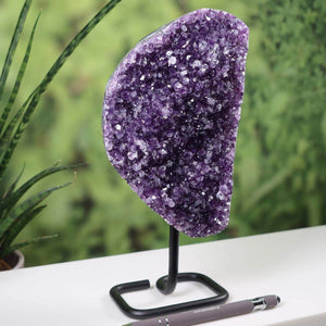 Deep purple Uruguay amethyst druse on a black metal stand, displayed on a white surface with green plants in the background.