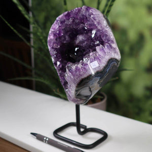 Deep purple Uruguay Amethyst Geode on metal stand displayed on a white table with a pen for scale, showcasing intense natural crystal formation.