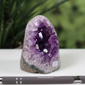 Intense purple Uruguay amethyst mini geode, unique piece number 27, displayed on a white surface with a pen and greenery in the background.