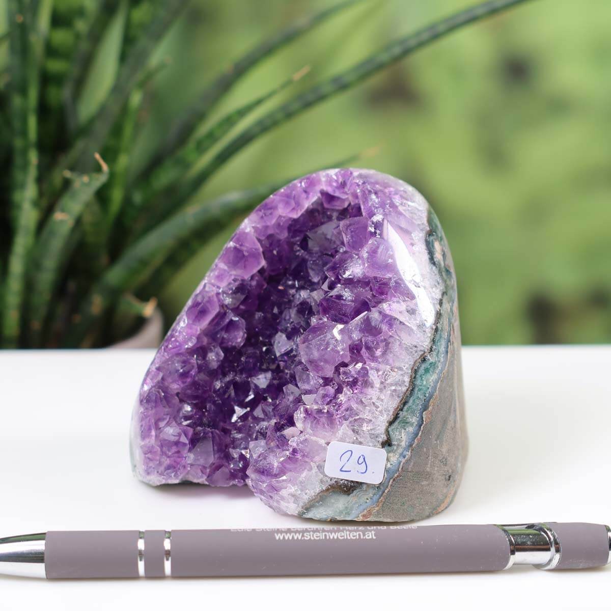 Intense purple Uruguay amethyst mini geode with natural crystal formation and label number 29 on a desk with a pen and plant background.