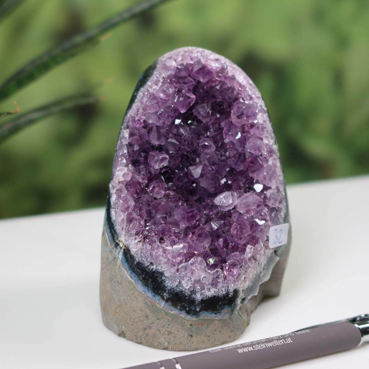 Uruguay Amethyst Mini Geode Number 32 with intense violet crystals displayed beside a pen on a desk with green plant background.