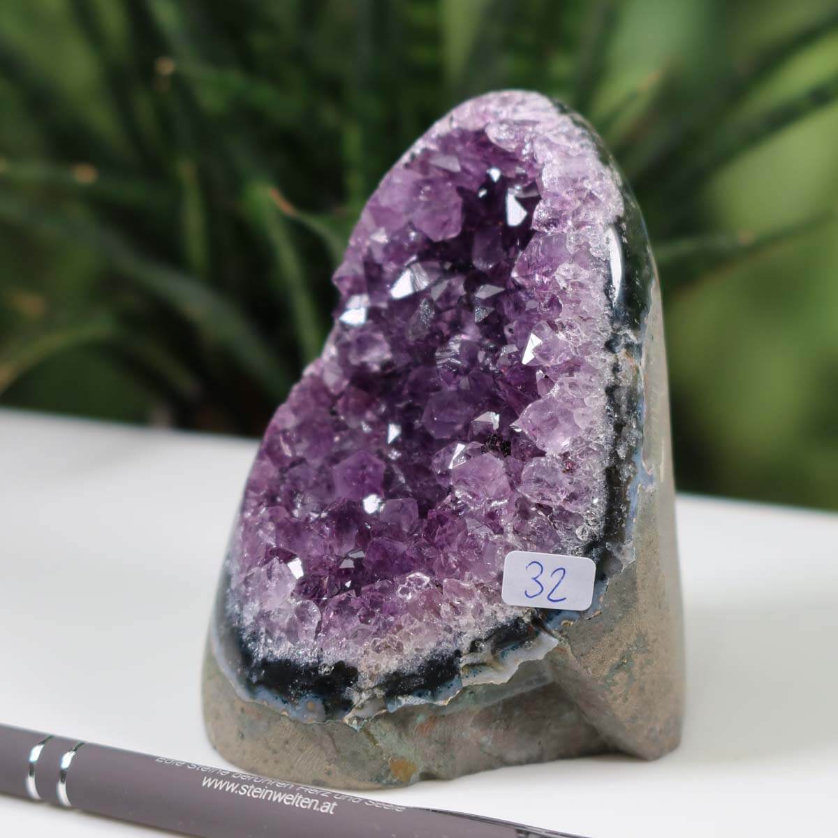 Uruguay Amethyst Geode Unikat NR 32 displayed on table, rich dark purple crystals inside gray rock, labeled with number "32", with a pen and plant background.