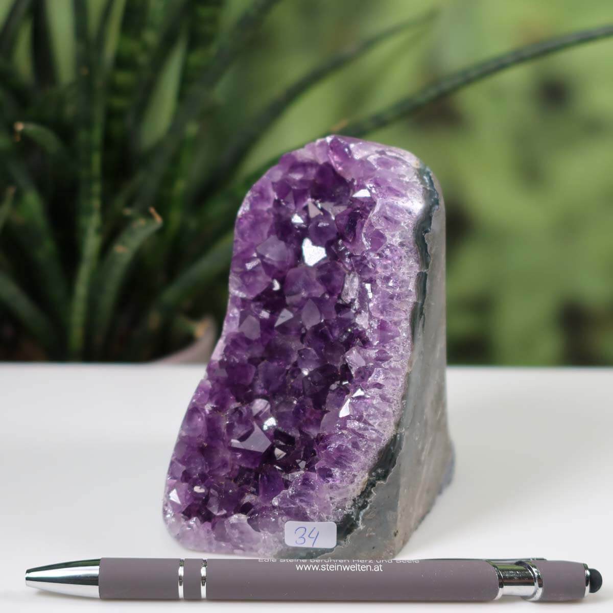 Uruguay Amethyst mini geode number 34 with intense violet crystals displayed beside a pen on a desk, natural green plant background.