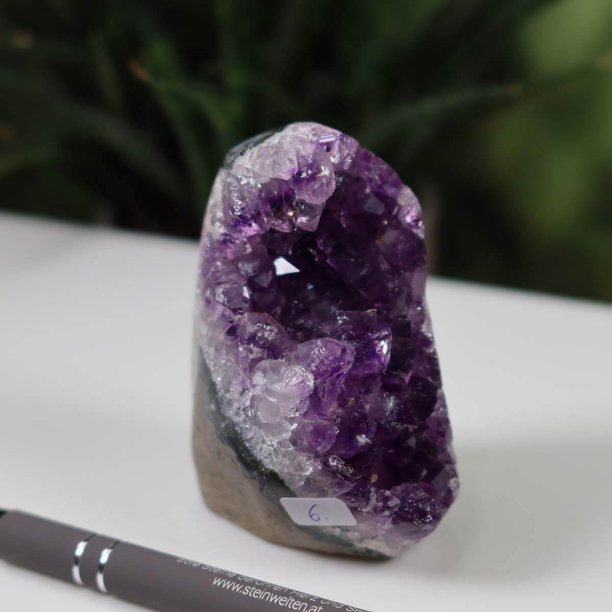 Intense violet Uruguay Amethyst mini geode, showing natural crystal patterns, labeled 'Unikat NR 6' on white surface with pen for scale.