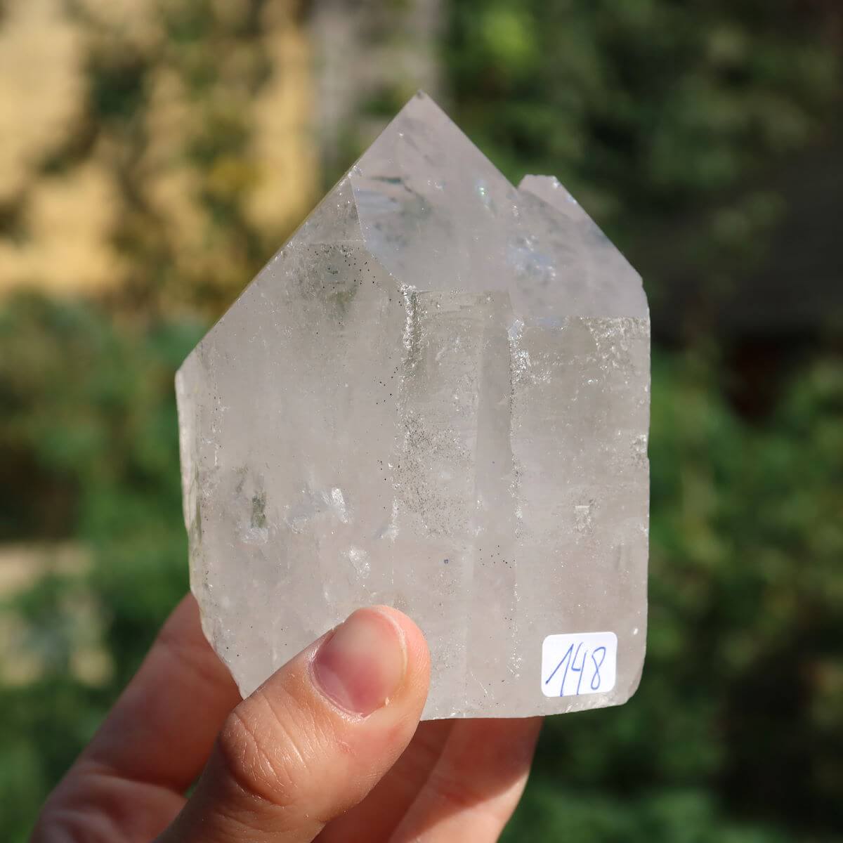 Hand holding polished clear quartz crystal point with facets, identified as Unikat Nr. 148, against a blurred natural background.