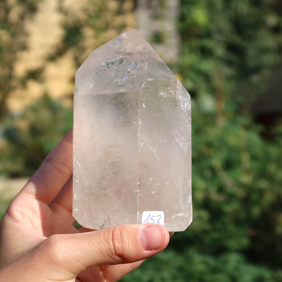 Hand holding a polished quartz crystal point from Brazil, measuring 10x7 cm, against a natural outdoor background.