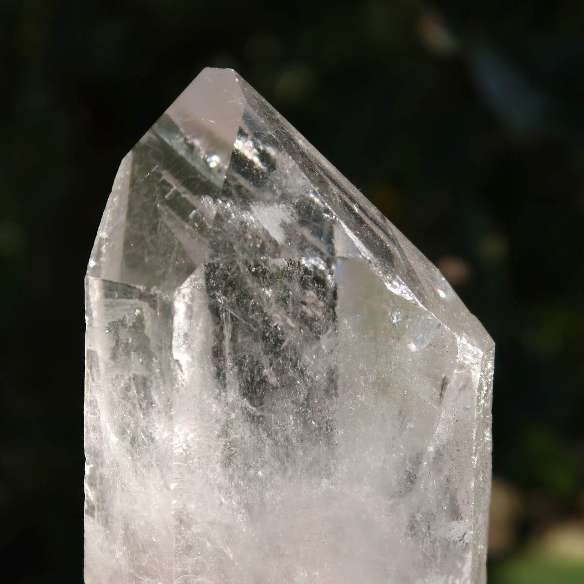 Hand holding a polished clear quartz crystal point, Bergkristall, against a blurred natural background, highlighting its clarity and facets.