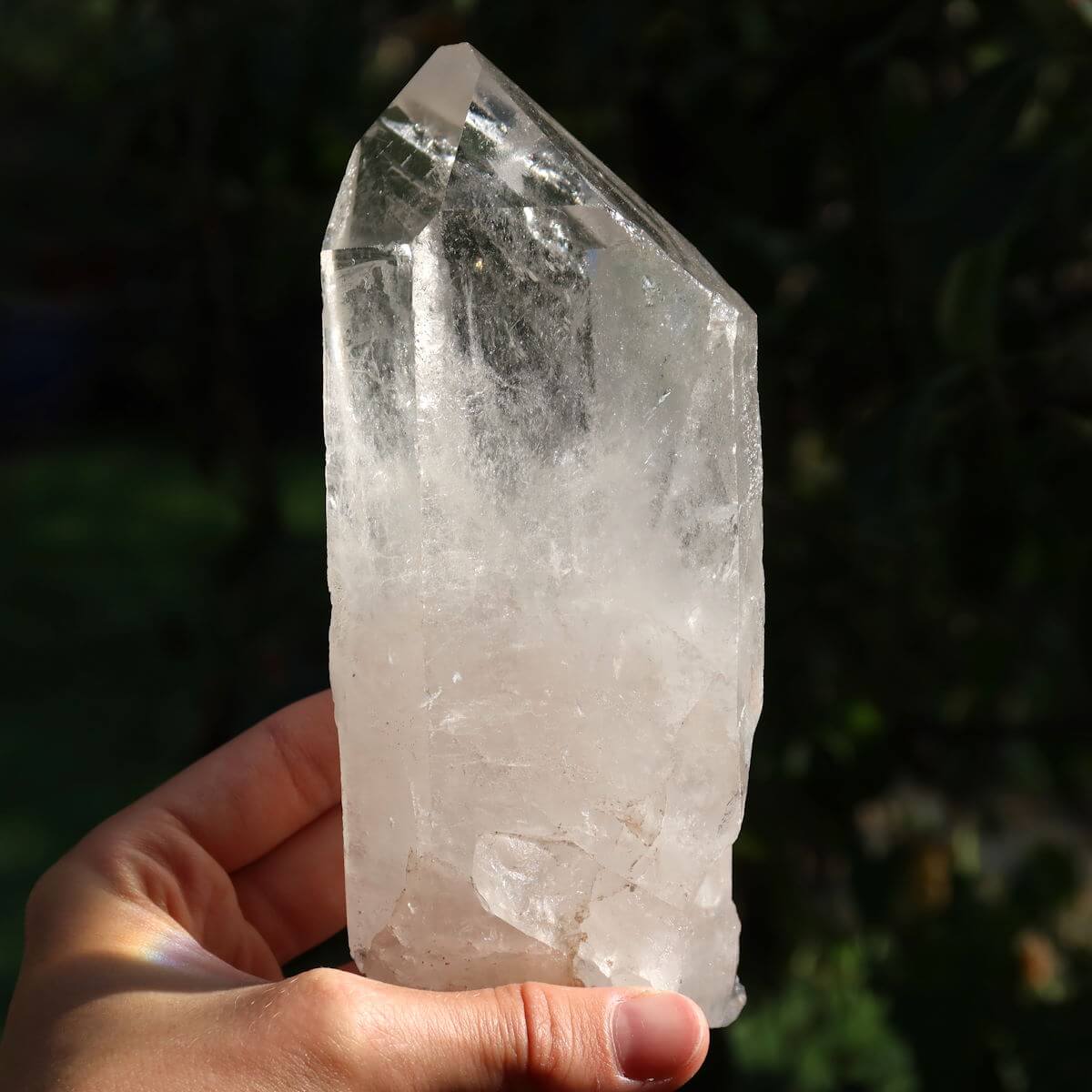 Hand holding a polished clear quartz crystal point, Bergkristall, against a blurred natural background, highlighting its clarity and facets.